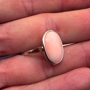 K&ouml;nnte beinhalten: Silberring mit einem ovalen, hellrosa Edelstein. Der Edelstein ist in einem schlichten Silberband gefasst. Das Design des Rings ist elegant und der Hintergrund ist unscharf.
