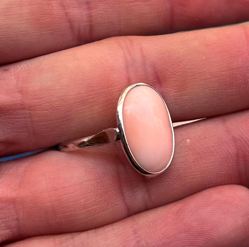 K&ouml;nnte beinhalten: Silberring mit einem ovalen, hellrosa Edelstein. Der Edelstein ist in einem schlichten Silberband gefasst. Das Design des Rings ist elegant und der Hintergrund ist unscharf.