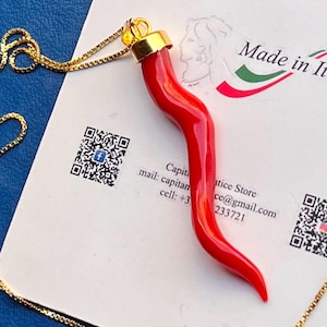 Op de afbeelding: Een felrode, gebogen hoornvormige hanger met een goudkleurige metalen dop, hangend aan een fijne goudkleurige boxketting. De hanger rust op een witte kaart met de tekst Made in It, een Facebook QR-code en contactgegevens.