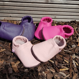 May include: Three pairs of soft leather baby shoes in purple, hot pink, and light pink. Each pair features a simple slip-on design with a rounded toe, a flexible sole, and a small white fabric tag on the side. The shoes have a textured grain finish.