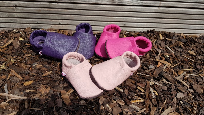 May include: Three pairs of soft leather baby shoes in purple, hot pink, and light pink. Each pair features a simple slip-on design with a rounded toe, a flexible sole, and a small white fabric tag on the side. The shoes have a textured grain finish.