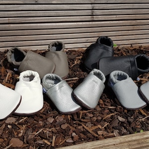 May include: A collection of five pairs of soft-soled baby shoes in various colors, including white, metallic silver, light gray, taupe, and black. Each pair features a rounded toe, a slip-on design with an elasticized ankle opening, and a textured finish.