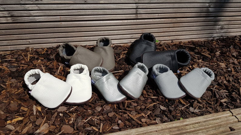 May include: A collection of five pairs of soft-soled baby shoes in various colors, including white, metallic silver, light gray, taupe, and black. Each pair features a rounded toe, a slip-on design with an elasticized ankle opening, and a textured finish.