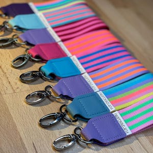 Könnte beinhalten: Eine Sammlung farbenfroher, gestreifter Taschenriemen mit silberfarbenen Verschlüssen. Die Riemen weisen eine Vielzahl lebendiger Farben auf, darunter Pink, Orange, Blau und Lila. Jeder Riemen hat ein Lederende mit dem Text "FUTZECLEIN".