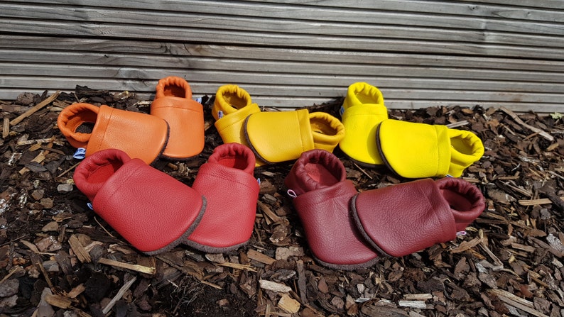 May include: Four pairs of soft leather baby slippers in vibrant colors including orange, yellow, bright red, and deep maroon. Each pair features a rounded toe, a flexible sole, and an elasticized ankle opening for a secure fit on small feet.