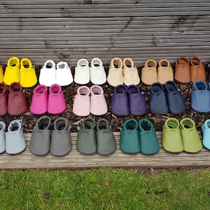 May include: A collection of soft leather baby slippers arranged in rows, featuring a wide variety of solid colors including orange, yellow, white, beige, brown, black, red, burgundy, pink, purple, navy, blue, silver, gray, olive, green, and teal.