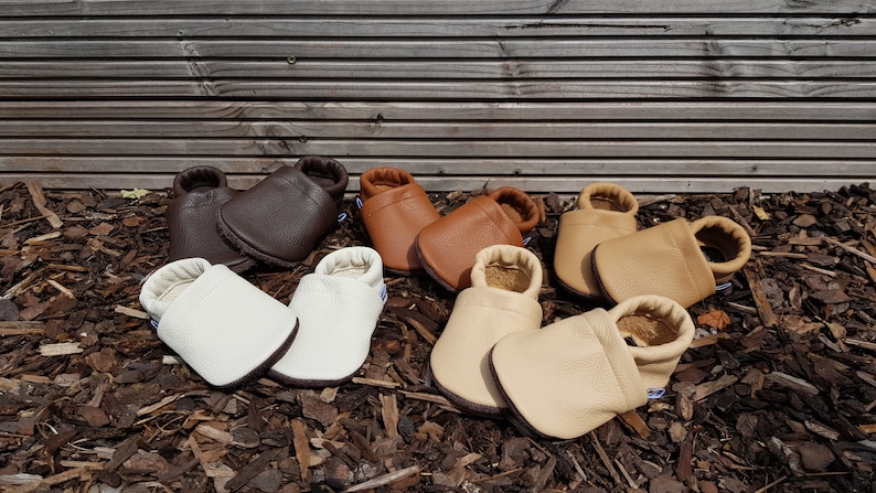 May include: Four pairs of soft-soled baby shoes in various shades of brown, tan, cream, and white. Each pair features a textured leather-like finish, a rounded toe, and an elasticized ankle opening for a secure fit on the feet.