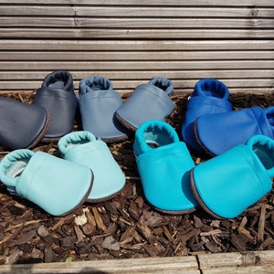 May include: A collection of four pairs of soft leather baby shoes in various shades of blue, including navy, slate blue, light turquoise, and bright cyan. Each pair features a rounded toe, a slip-on design, and a flexible sole for comfort.