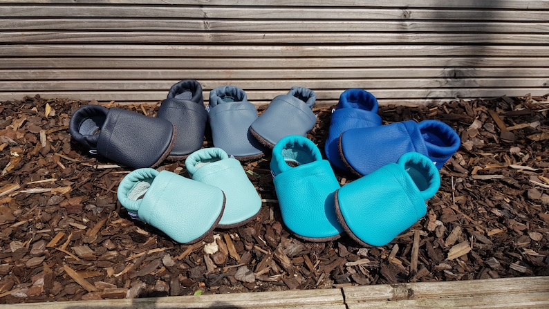 May include: A collection of four pairs of soft leather baby shoes in various shades of blue, including navy, slate blue, light turquoise, and bright cyan. Each pair features a rounded toe, a slip-on design, and a flexible sole for comfort.