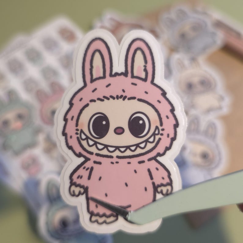 K&ouml;nnte beinhalten: Ein rosa Aufkleber mit einem Cartoon-Monster in einem Hasenkost&uuml;m. Das Monster hat gro&szlig;e schwarze Augen, ein breites Grinsen und einen pelzigen rosa K&ouml;rper. Weitere Aufkleber sind im Hintergrund zu sehen.