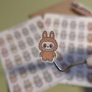 May include: A brown sticker featuring a cartoon character in a furry bunny costume with a wide, toothy grin and large, round eyes. The sticker is held by a dental tool, with a sheet of similar stickers in the background.