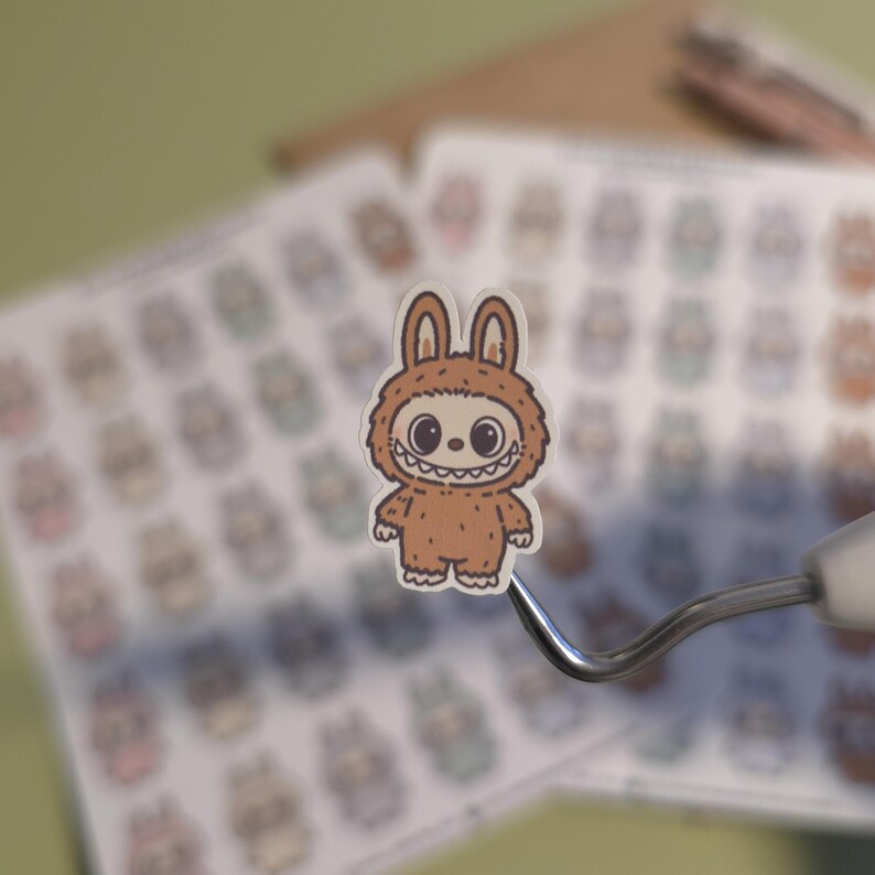 May include: A brown sticker featuring a cartoon character in a furry bunny costume with a wide, toothy grin and large, round eyes. The sticker is held by a dental tool, with a sheet of similar stickers in the background.