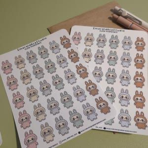 May include: Two sheets of stickers featuring cute monster designs. Each sticker showcases a bunny-like creature in various pastel colors, including pink, brown, and gray. The stickers are printed on white paper, with the text "EmeraldAceStudio cute monsters" at the top.