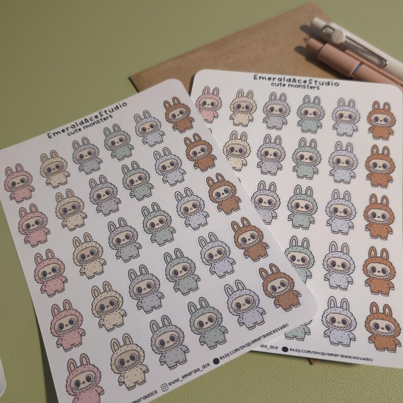 May include: Two sheets of stickers featuring cute monster designs. Each sticker showcases a bunny-like creature in various pastel colors, including pink, brown, and gray. The stickers are printed on white paper, with the text "EmeraldAceStudio cute monsters" at the top.
