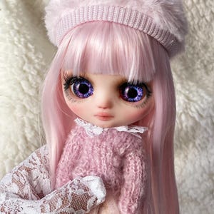 Könnte beinhalten: Eine rosa Puppe mit langen rosa Haaren, die einen rosa Pullover und einen rosa Hasenohrhut trägt. Die Puppe hat große, ausdrucksstarke Augen mit violetten Iris und langen Wimpern. Das Outfit der Puppe umfasst einen weißen Spitzenkragen.