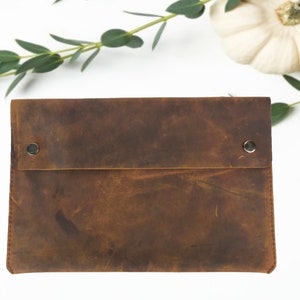 May include: A brown leather envelope clutch with two silver snaps. The clutch has a distressed look and is made of a soft, supple leather.