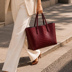 May include: A beige blazer worn over a matching cream-colored tank top and high-waisted trousers, paired with a dark brown leather belt, pointed-toe heels, and a large, deep burgundy tote bag with a textured, embossed crocodile-skin pattern.