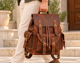 May include: A brown leather backpack featuring a top flap with two buckle straps, a drawstring closure, and three exterior pockets with buckle fastenings. The bag includes adjustable shoulder straps and antique brass-toned metal hardware accents.