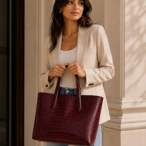 May include: A dark burgundy tote bag with a textured crocodile-embossed pattern, featuring two sturdy top handles, a rectangular silhouette, and gold-toned metal feet at the base for stability, presented in a structured and polished design.