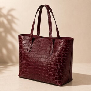 May include: A deep burgundy tote bag featuring a textured crocodile skin pattern. The bag has two vertical handles attached to the top edge, a rectangular base, and a structured silhouette with a smooth interior lining visible at the opening.