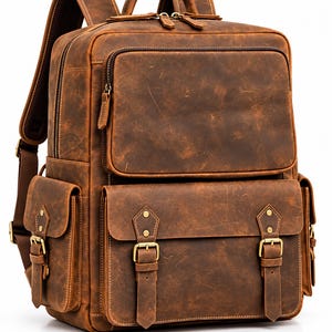 Könnte beinhalten: Ein brauner Lederrucksack mit einem großen Hauptfach mit Reißverschluss, einer zusätzlichen Reißverschlusstasche vorne und einer unteren Vordertasche, die mit zwei Schnallenriemen und Messingbeschlägen gesichert ist. Zwei Seitentaschen.