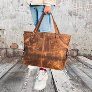 May include: A brown leather tote bag with two handles and adjustable straps. The bag has a distressed finish and metal buckles. The bag is held by a person wearing blue jeans and white sneakers.