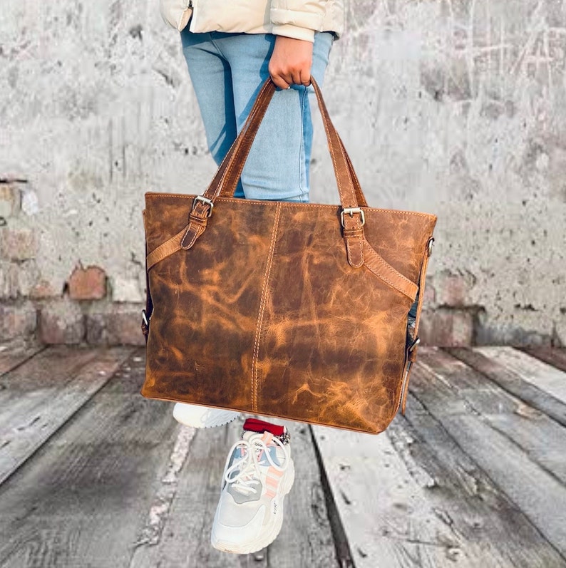 May include: A brown leather tote bag with two handles and adjustable straps. The bag has a distressed finish and metal buckles. The bag is held by a person wearing blue jeans and white sneakers.