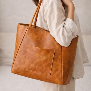 May include: A large, rectangular brown tote bag with a front pocket. The bag has two long handles and is made of a textured, leather-like material. The bag is being held over a person's shoulder.