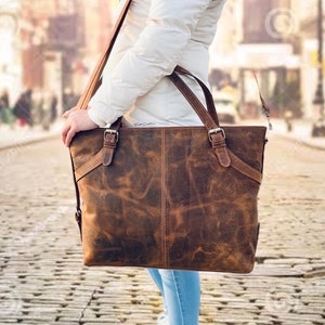 May include: A brown leather tote bag with a distressed finish. The bag has two top handles and a shoulder strap, secured with silver-colored buckles. The bag is carried by a person wearing a white jacket and blue jeans.