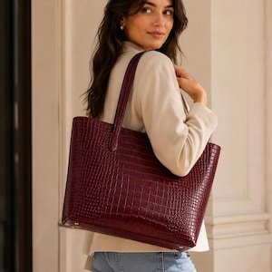 May include: A deep burgundy tote bag featuring a textured crocodile-embossed pattern. The bag has a structured rectangular shape, two vertical shoulder straps, and visible gold-toned metal hardware at the base corners for added durability.