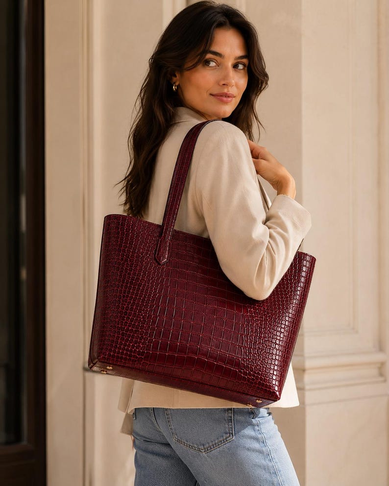 May include: A deep burgundy tote bag featuring a textured crocodile-embossed pattern. The bag has a structured rectangular shape, two vertical shoulder straps, and visible gold-toned metal hardware at the base corners for added durability.