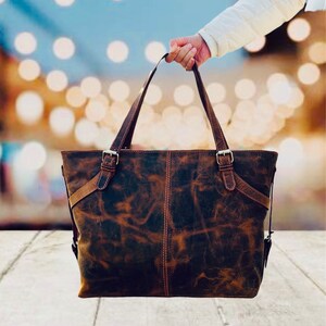 May include: A distressed brown leather tote bag with two top handles and adjustable shoulder straps. The bag features metal buckles and stitching details. The leather has a mottled appearance, with variations in color and texture.