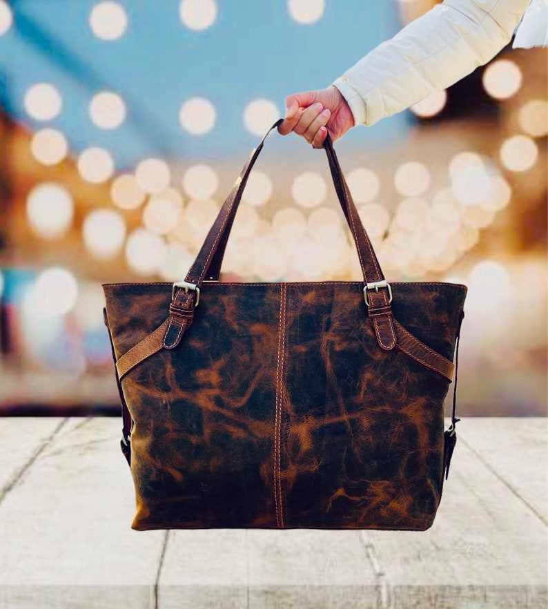 May include: A distressed brown leather tote bag with two top handles and adjustable shoulder straps. The bag features metal buckles and stitching details. The leather has a mottled appearance, with variations in color and texture.