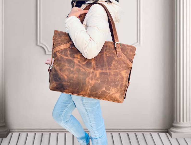 May include: A large, brown leather tote bag with a distressed finish. The bag has two top handles and a shoulder strap. The bag is rectangular and has a buckle closure. The bag is carried by a person wearing a white jacket and blue jeans.