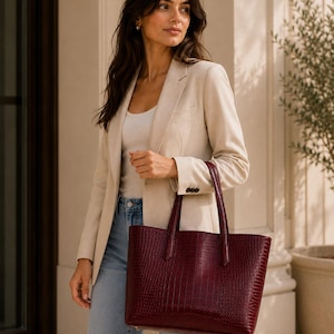 May include: A deep burgundy tote bag with a textured crocodile-embossed finish, featuring two sturdy top handles and a structured rectangular shape. The bag is crafted with a polished exterior and includes visible metal hardware at the base corners.