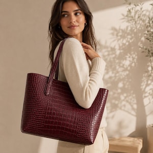 May include: A deep burgundy tote bag featuring a textured crocodile-embossed pattern. The bag has a structured rectangular shape with two vertical shoulder straps attached to the top edge, providing a clean and functional design for daily use.