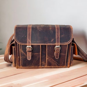 Puede incluir: Una bolsa de cámara de cuero marrón con dos correas ajustables y dos bolsillos delanteros. La bolsa tiene un aspecto desgastado y está hecha de cuero de alta calidad.