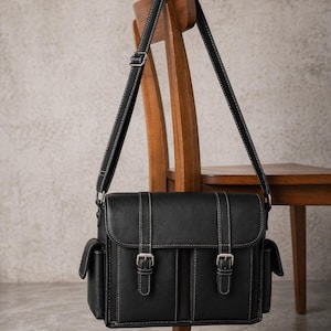 May include: Black messenger bag with white stitching and silver buckles. The bag has a flap closure and two front pockets. The bag is hanging on a wooden chair. The bag's strap is also black with white stitching.