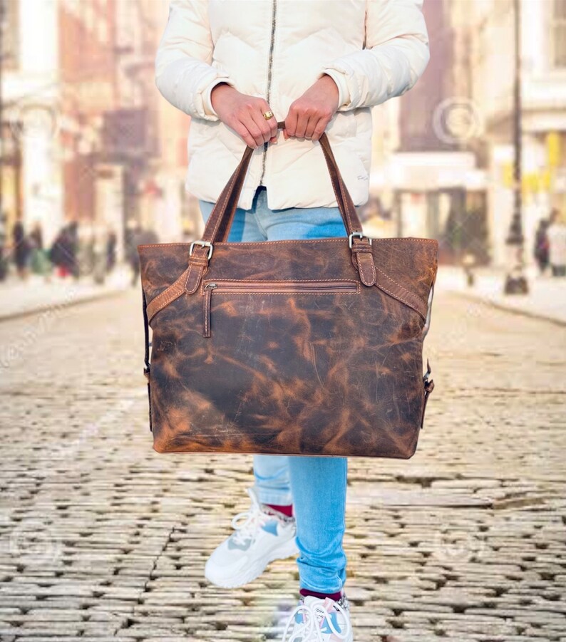 May include: A distressed brown leather tote bag with two handles and a zippered pocket. The bag has a rugged, textured appearance and is held by a person wearing a white jacket and blue jeans. The bag features metal buckles and a top zipper.