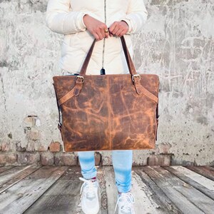 May include: A large, brown leather tote bag with two top handles and adjustable side straps. The bag has a distressed finish, visible stitching, and metal buckles. The bag is held by a person wearing a white jacket and blue jeans.