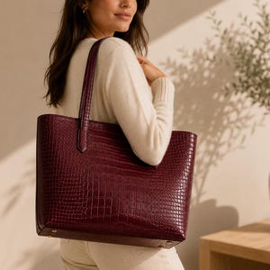 May include: A deep burgundy tote bag featuring a textured crocodile-embossed pattern. The bag has a structured rectangular shape with two vertical shoulder straps attached to the top edge and small metallic feet on the bottom base for stability.