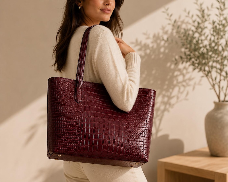 May include: A deep burgundy tote bag featuring a textured crocodile-embossed pattern. The bag has a structured rectangular shape with two vertical shoulder straps attached to the top edge and small metallic feet on the bottom base for stability.