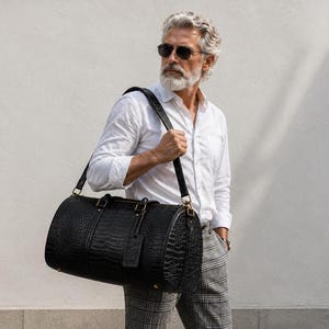 May include: A black cylindrical duffel bag featuring a textured crocodile-embossed pattern, gold-toned metal hardware, two top handles, and a detachable shoulder strap. The bag includes a matching luggage tag and a secure zipper closure.