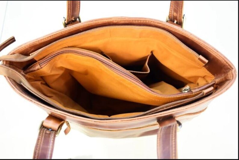 May include: Open view of a brown leather tote bag with multiple compartments. The bag has two handles and a shoulder strap. The interior is lined with a tan fabric and features a zippered pocket and several open pockets. The bag is made of a distressed leather.