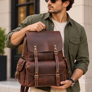 Puede incluir: Una mochila de cuero marrón con una solapa superior grande, dos correas verticales con hebilla y dos bolsillos laterales. La bolsa incluye herrajes de metal en tono latón, costuras visibles y un asa superior, con un diseño estructurado.