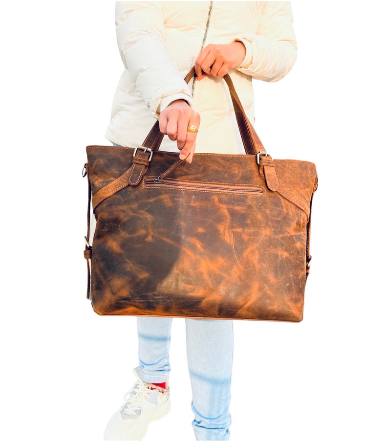 May include: A brown leather tote bag with a distressed finish. The bag has two top handles and a zippered pocket on the front. The bag is held by a person wearing a white jacket and blue jeans.