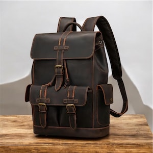 May include: A dark brown leather backpack with multiple pockets and adjustable straps. The backpack features a flap closure with buckles and decorative stitching. The bag is sitting on a wooden surface.