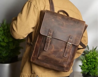 May include: A brown leather backpack with two adjustable buckles on the front flap. The backpack has a distressed look and is being worn by a person.
