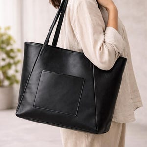May include: A black tote bag with a rectangular front pocket. The bag has two long handles and is made of a smooth material. The bag is being held by a person wearing a light-coloured shirt and trousers.