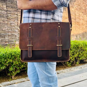 May include: A brown leather messenger bag with a flap closure and adjustable shoulder strap. The bag features two vertical straps with gold-colored buckles and decorative stitching. The bag is worn over the shoulder.